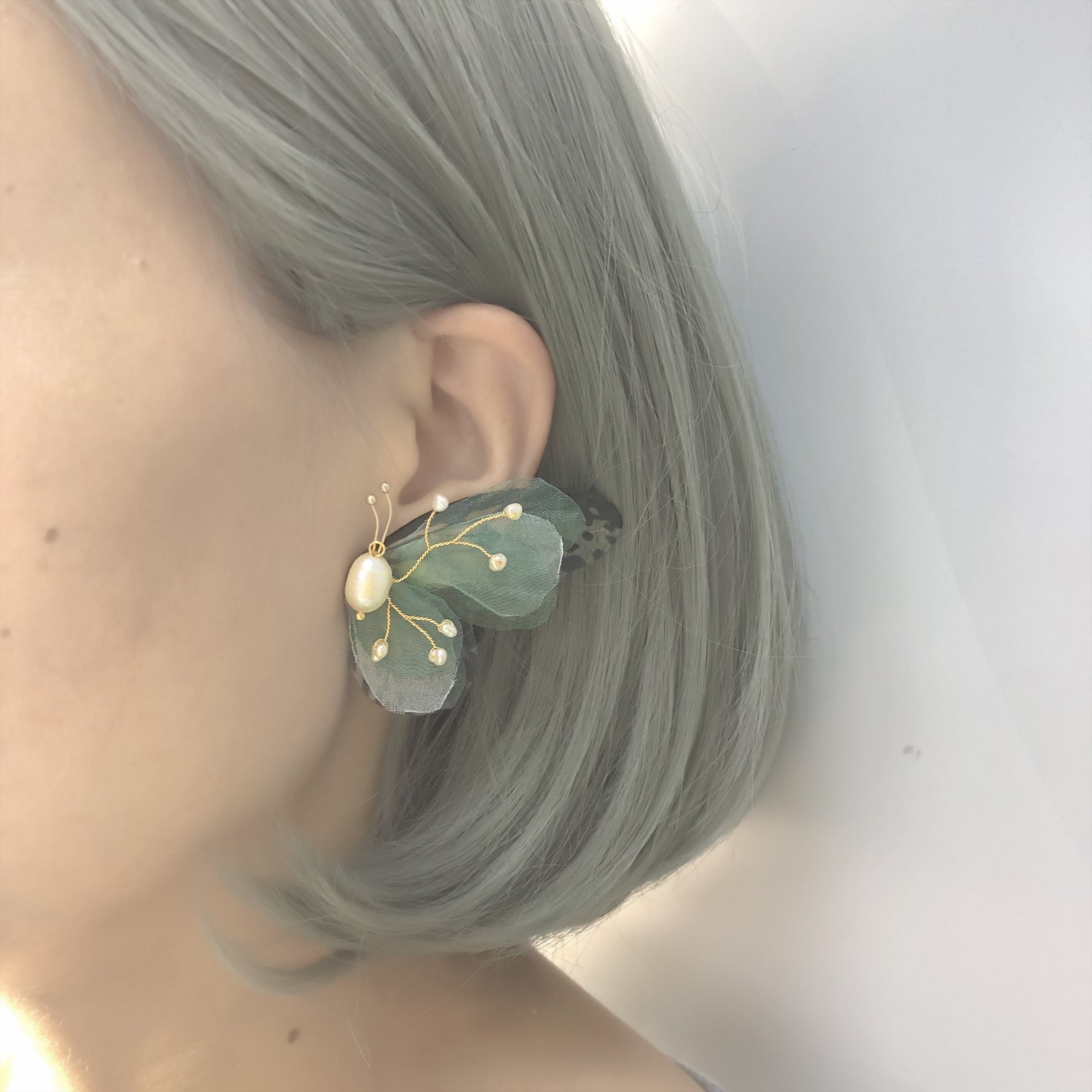 Close-up side profile of a woman wearing a large butterfly earring with green wings, freshwater pearls, and golden accents. The woman's hair is styled in a sleek, gray bob.