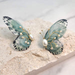 Delicately designed green butterfly earrings with soft translucent wings, freshwater pearls, and golden wire accents, photographed on a textured beige background.