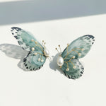 Green butterfly earrings with translucent wings, gold wire details, and pearl accents on white background.