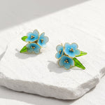 Floral earrings with blue flowers and green leaves on a white stone surface