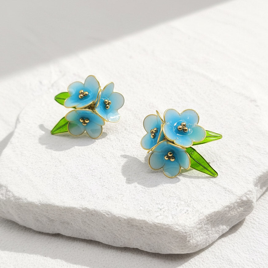 Floral earrings with blue flowers and green leaves on a white stone surface