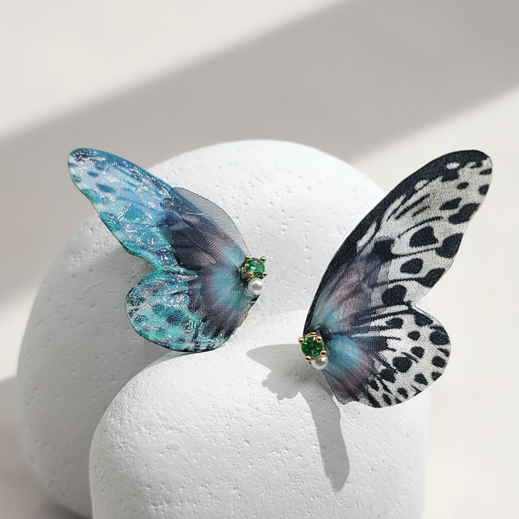 Blue and black butterfly wing earrings with green gemstone and pearl accents displayed on white stone.