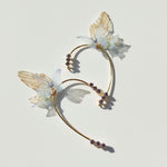 Gold butterfly ear cuffs with translucent wings, pearl accents, and purple crystals on light background.