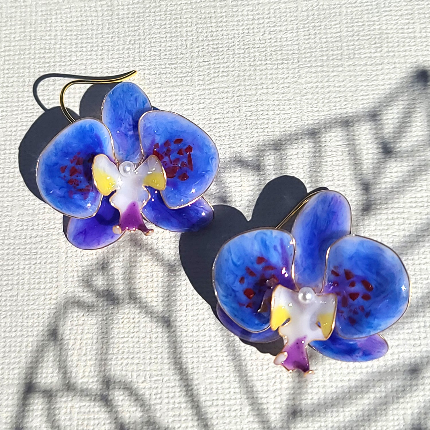 Artisan-crafted blue enamel orchid earrings with gold-plated hooks, displayed on textured canvas with natural shadow patterns