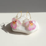 Hand-painted pink and white orchid flower earrings with pearl and crystal accents, displayed in soft sunlight.