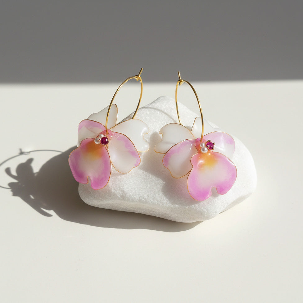 Hand-painted pink and white orchid flower earrings with pearl and crystal accents, displayed in soft sunlight.