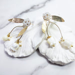 A pair of gold-plated stud earrings featuring hand-carved mother-of-pearl tulip drops, delicate freshwater pearls, and beaded glass floral studs with gold leaf accents, displayed on white textured petals.