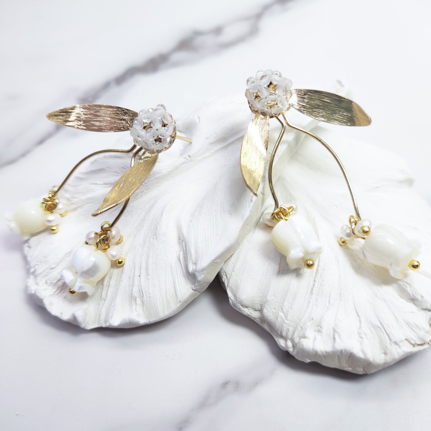 A pair of gold-plated stud earrings featuring hand-carved mother-of-pearl tulip drops, delicate freshwater pearls, and beaded glass floral studs with gold leaf accents, displayed on white textured petals.