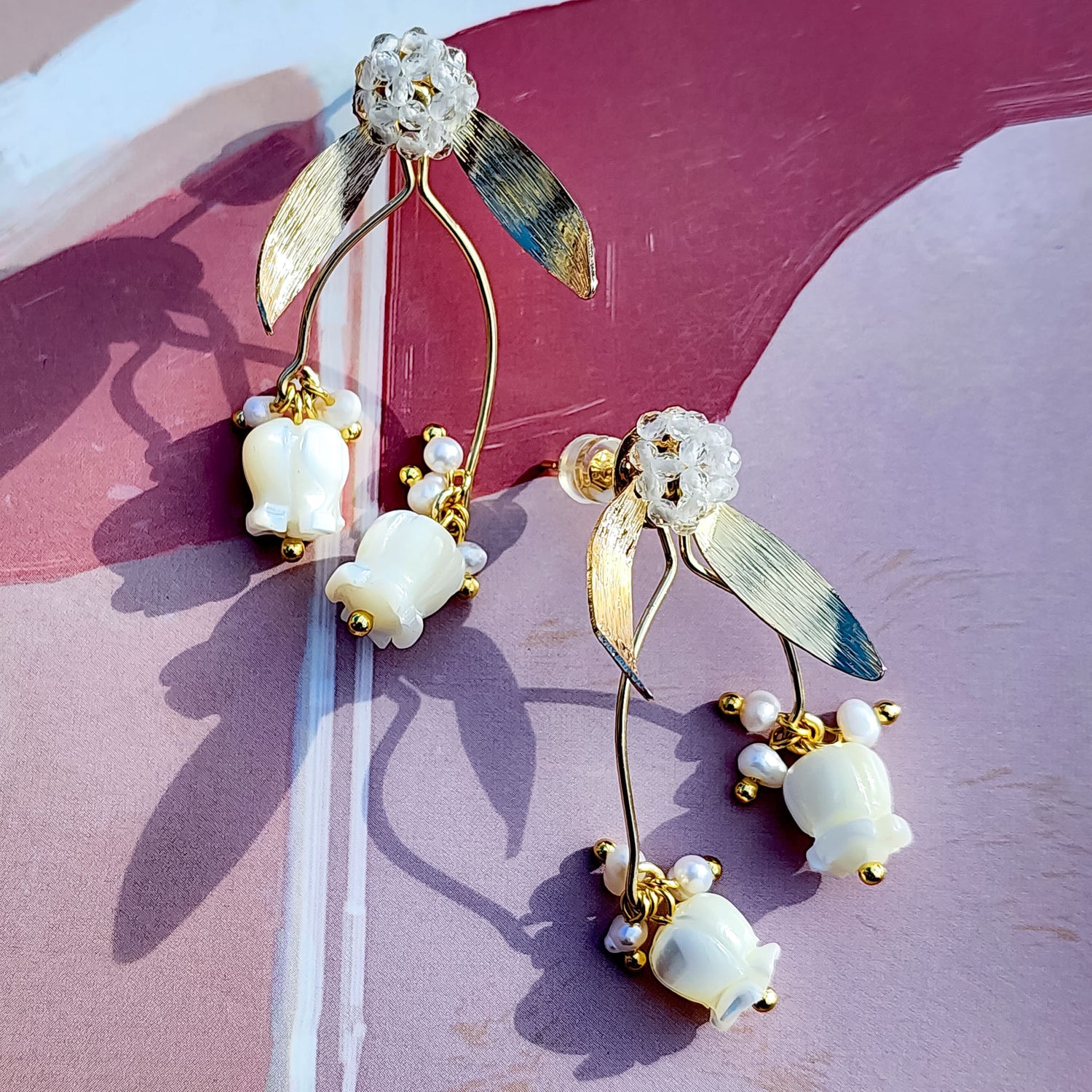 Gold-plated stud earrings featuring hand-carved mother-of-pearl tulip drops and freshwater pearl accents, suspended from beaded glass floral studs with metallic leaf details, displayed on a pink and mauve backdrop with natural shadows.