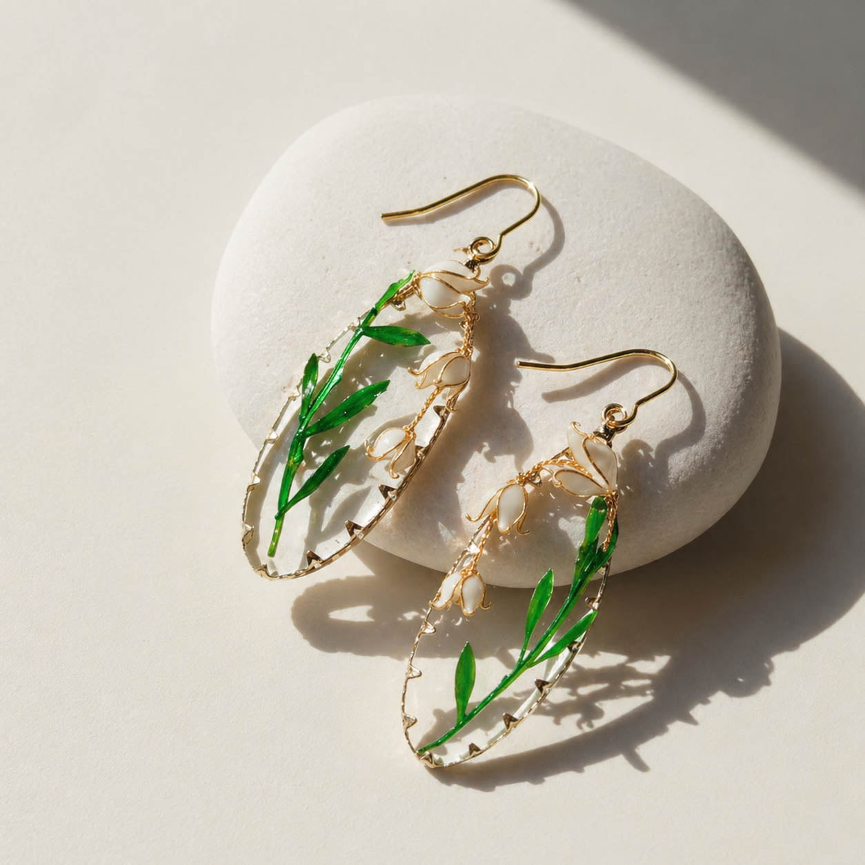 Gold and green leaf-shaped earrings with lily of the valley flowers on a white stone surface