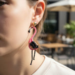 Close-up of a person wearing pink and black beaded earrings with a blurred outdoor background.