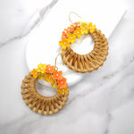 Handwoven wicker circle earrings with vibrant orange and yellow resin hydrangea flowers, displayed on a white marble background.