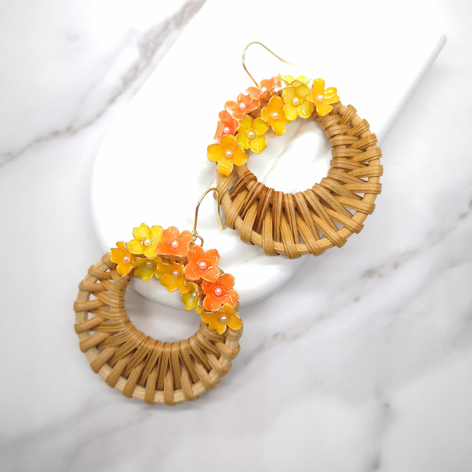 Handwoven wicker circle earrings with vibrant orange and yellow resin hydrangea flowers, displayed on a white marble background.