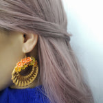 Model wearing large handwoven wicker hoop earrings adorned with orange and yellow hand-painted resin hydrangea flowers, styled with long mauve hair and a blue sweater.