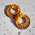 Handwoven wicker hoop earrings with yellow and orange handpainted hydrangea flowers, displayed on terrazzo surface in natural sunlight.