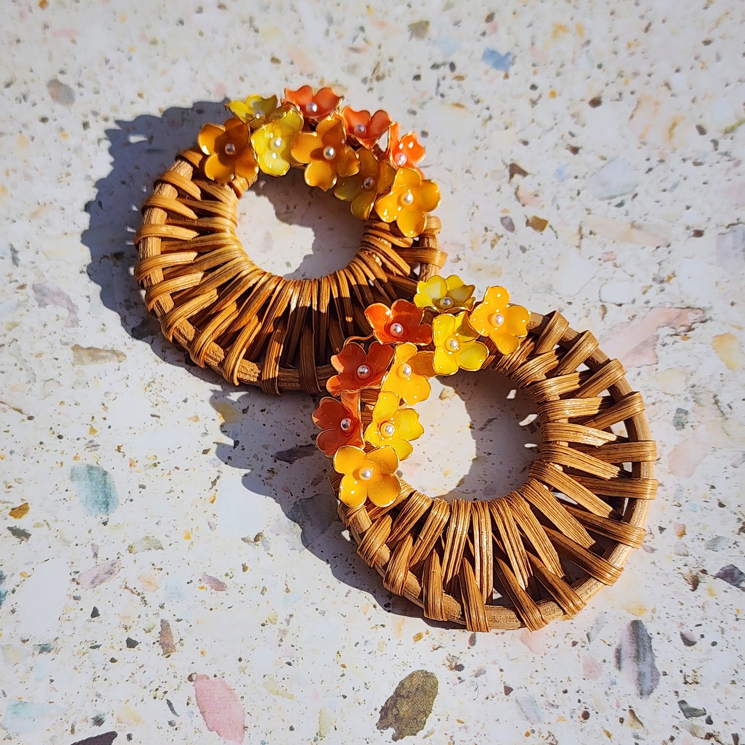 Handwoven wicker hoop earrings with yellow and orange handpainted hydrangea flowers, displayed on terrazzo surface in natural sunlight.