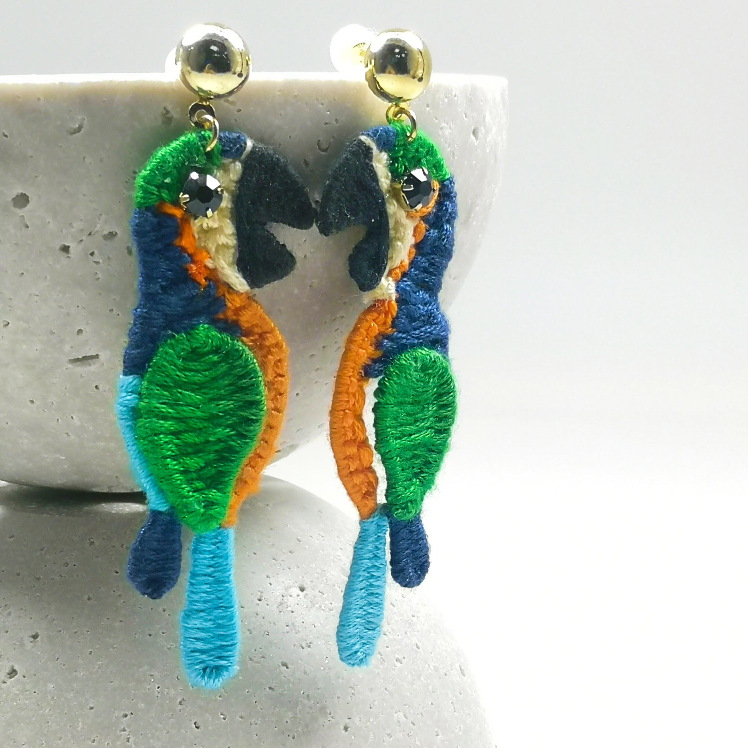 Colorful embroidered parrot earrings with green, blue, and orange thread, featuring black crystal eyes and gold-plated studs, displayed on a textured ceramic stand.