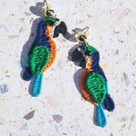 Handwoven tropical parrot embroidered earrings in vibrant blue, green, and orange thread with gold-plated studs, photographed on terrazzo background.