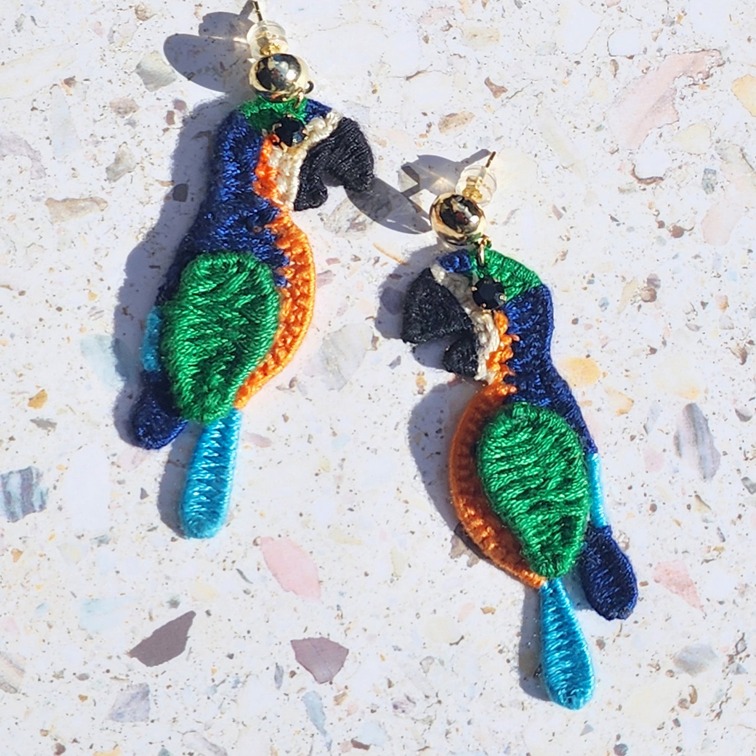 Handwoven tropical parrot embroidered earrings in vibrant blue, green, and orange thread with gold-plated studs, photographed on terrazzo background.