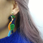 Colorful embroidered parrot earring with green, orange, and blue thread shown worn on model, featuring a gold-plated stud and statement tropical bird design.