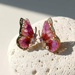 February birthstone butterfly-shaped earrings with pink wings and gold accents on a textured stone surface.