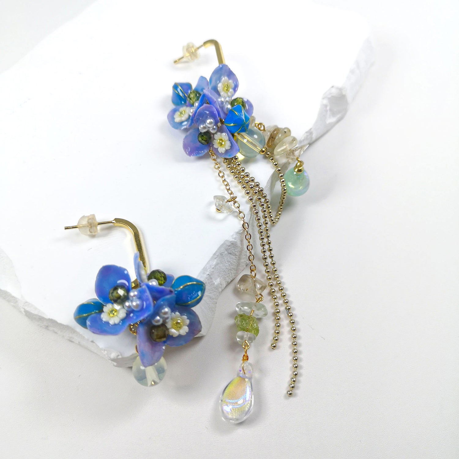 Asymmetrical gold-plated dangle earrings featuring real blue hydrangea flowers, hand-painted resin petals, semi-precious stone chips, and a teardrop glass charm.