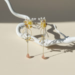 Delicate earrings with floral designs and pink stones on a white branch against a beige background