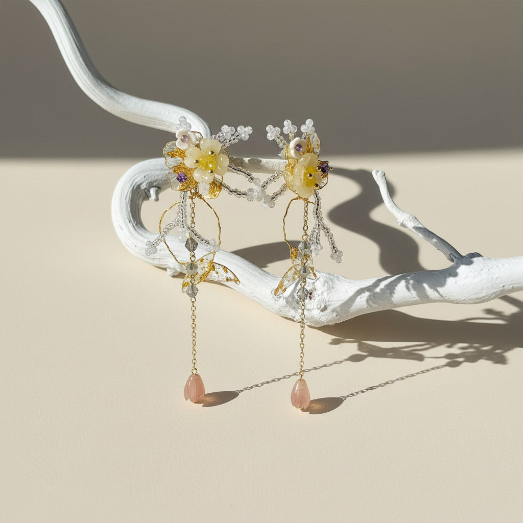 Delicate earrings with floral designs and pink stones on a white branch against a beige background