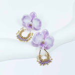 Pair of lavender orchid earrings with pearl centers and gold beaded hoops, featuring purple crystal accents, displayed on a white curved surface.