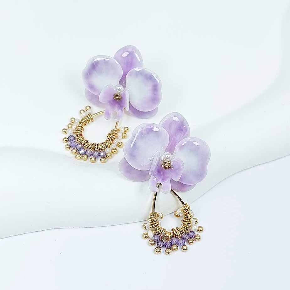 Pair of lavender orchid earrings with pearl centers and gold beaded hoops, featuring purple crystal accents, displayed on a white curved surface.