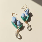 Blue tulip-shaped earrings with green leaves and clear teardrop pendants on a beige background