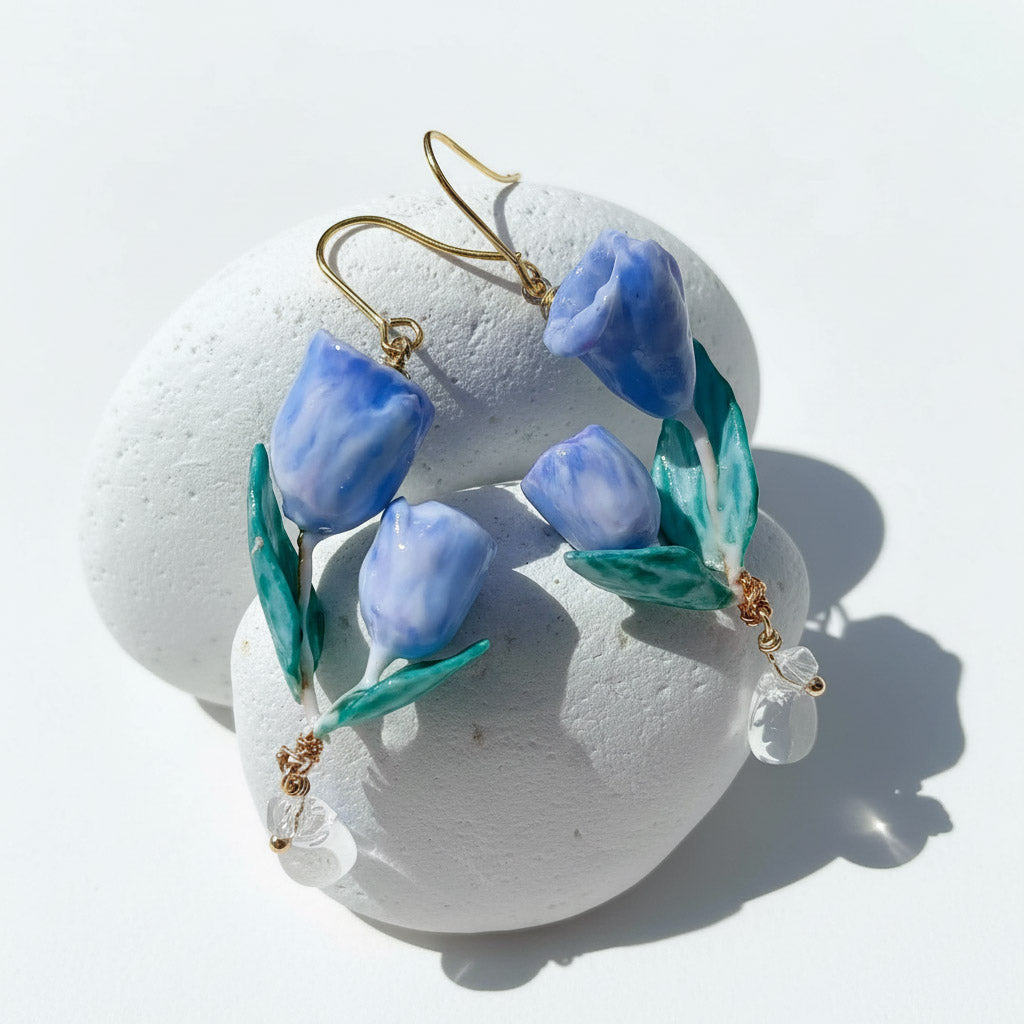 Handcrafted blue tulip flower earrings with green leaves and crystal drops, resin floral jewelry on white stone background.