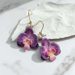 Purple flower earrings with gold hooks on a white stone surface