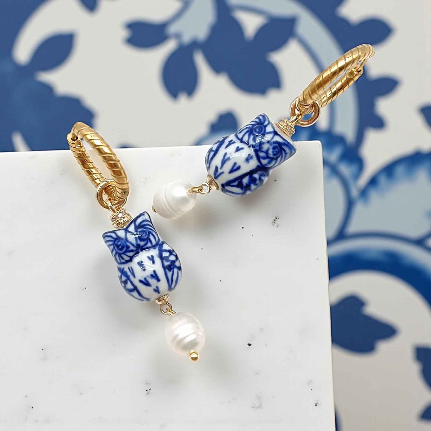 Earrings with blue and white ceramic beads and gold accents on a white surface with a blue floral pattern in the background.