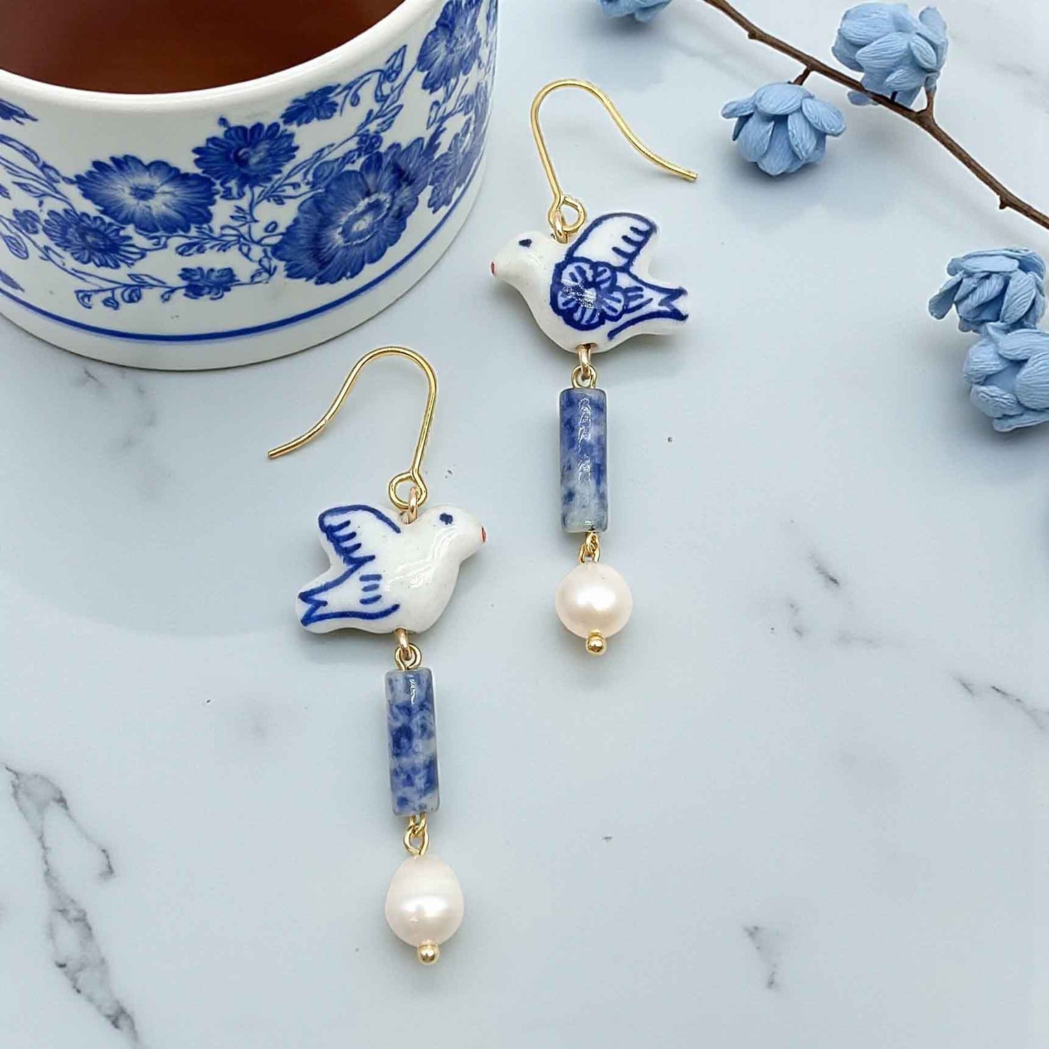Ceramic dove earrings with blue and white design on a marble surface with a floral teacup.