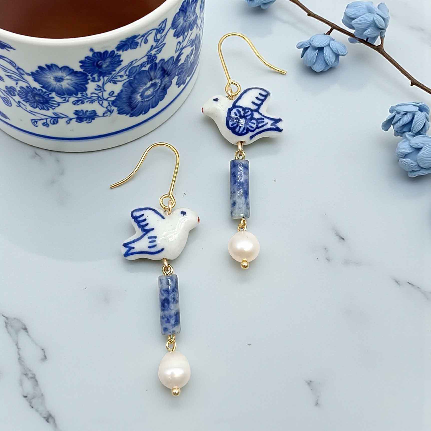 Ceramic dove earrings with blue and white design on a marble surface with a floral teacup.