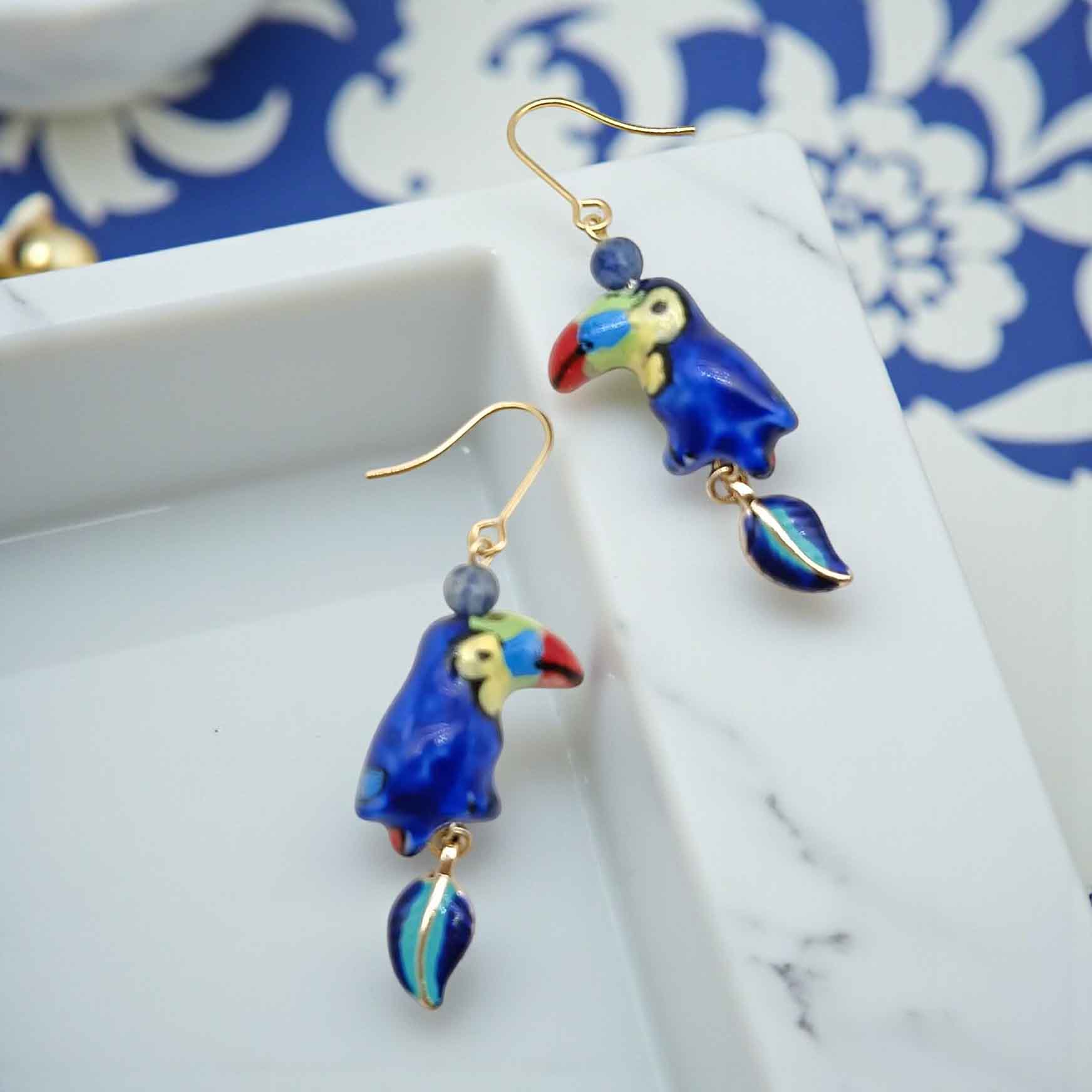 Blue parrot-shaped earrings on a white marble surface with a blue floral patterned fabric in the background.