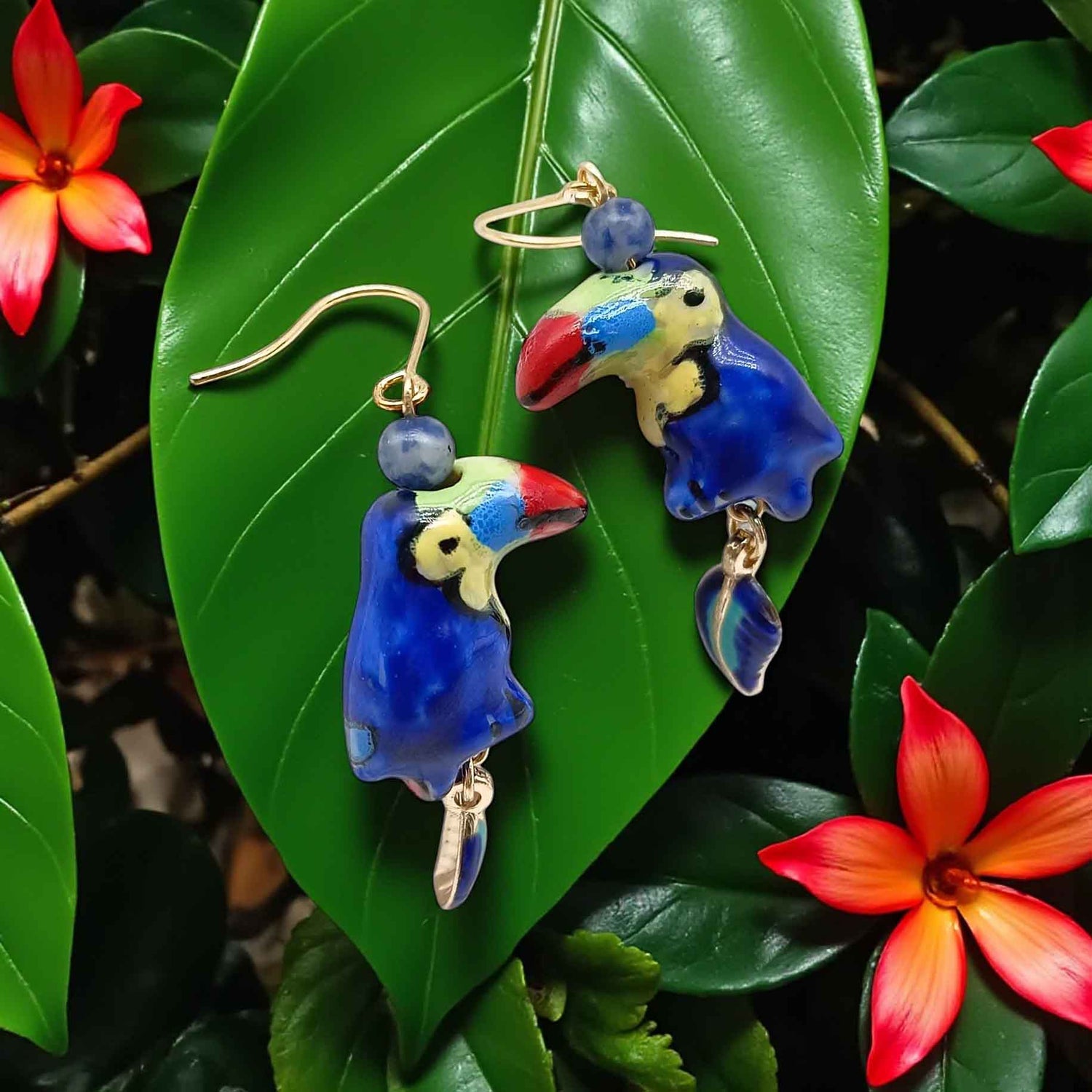 Blue parrot-shaped earrings on a green leaf with red flowers in the background