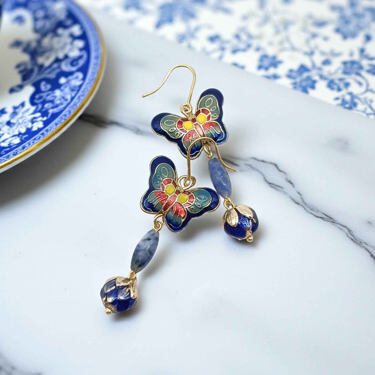 Blue cloisonné butterfly earrings with lapis stone drop on marble surface, artistic jewelry styling.