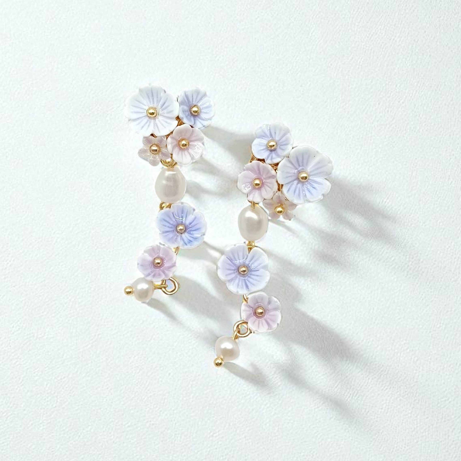 Close-up of white and lilac floral pearl drop earrings on soft white surface, gold-plated detailing.