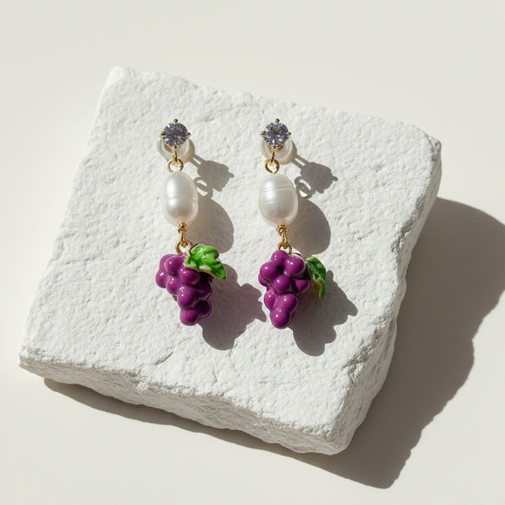 Hand-painted enamel grape and pearl drop earrings on white stone background, sunlight and shadow.