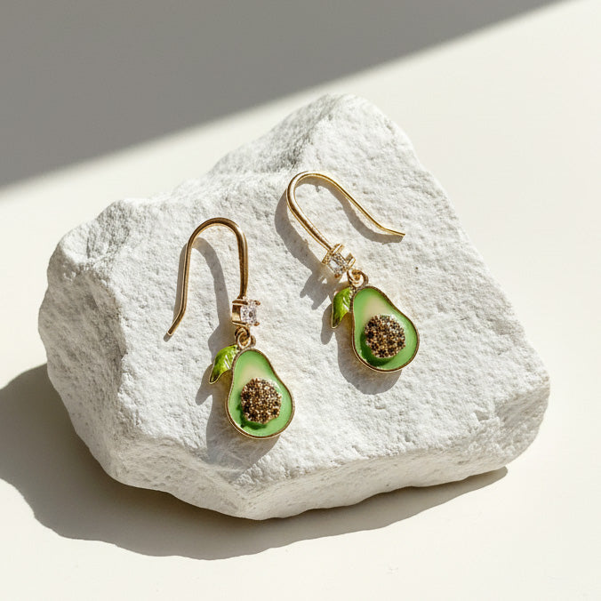 Close-up of enamel avocado earrings with CZ sparkle and gold-plated hooks on white stone.