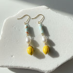 Close-up of gold-plated lemon enamel earrings with pearl and aqua stone on white surface.