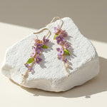 Purple and green wisteria floral drop earrings displayed on textured stone.