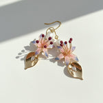 Close-up of pink and lilac hand-painted flower earrings with pearl and gold leaf charm under soft natural light.