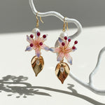 Hanging view of gold-plated pink lily floral earrings with pearl accents and CZ leaf details, sunlight and shadows.