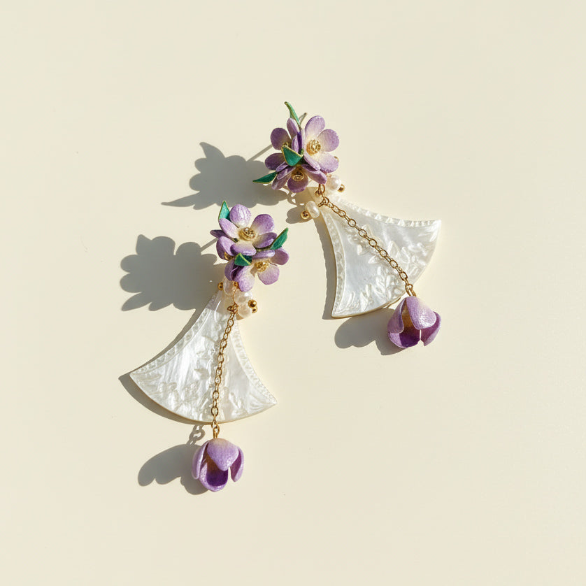 Iridescent shell and lilac flower earrings displayed on beige background.