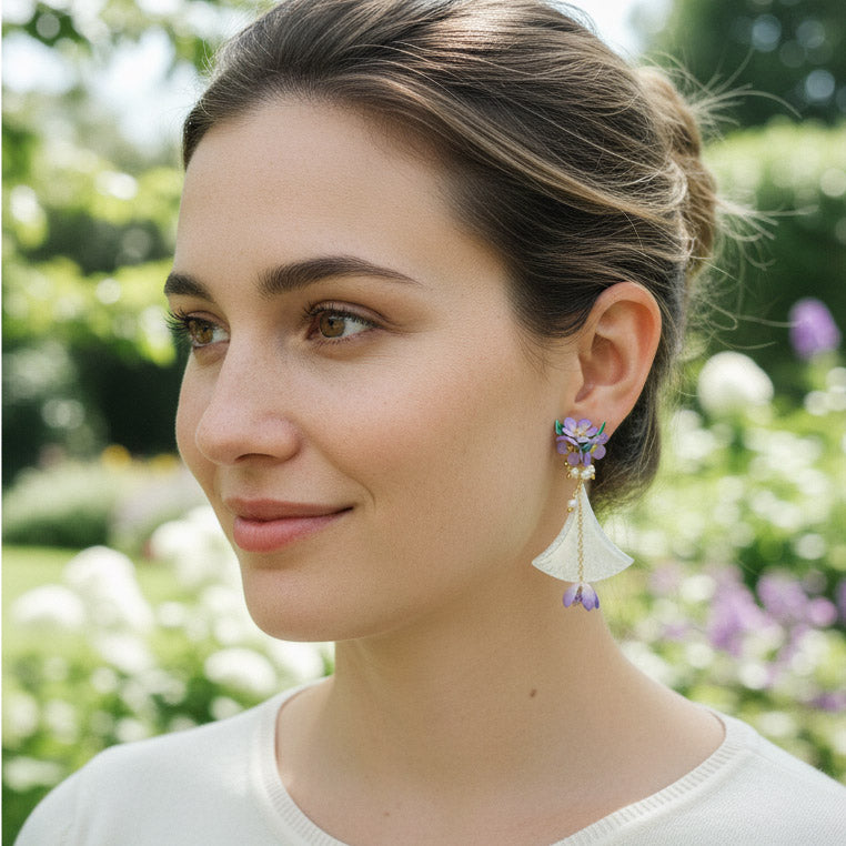 Model wearing hand-painted lilac flower and shell fan earrings in garden light.