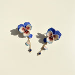 Hand-painted blue and white pansy flower earrings with gold chains and blue bead drops on cream background.