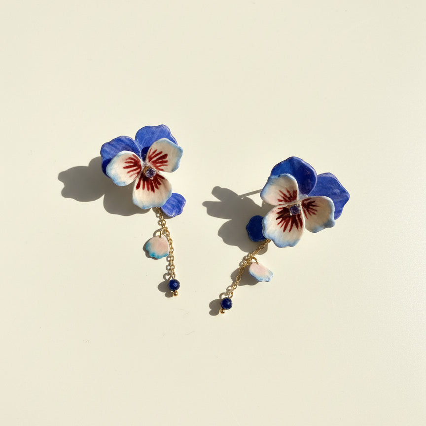 Hand-painted blue and white pansy flower earrings with gold chains and blue bead drops on cream background.